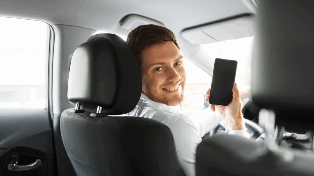 Mann sitzt im Auto und zeigt lächelnd auf sein Smartphone – Symbolbild für die einfache Online-Abfrage von Flensburg Punkten Das Bild zeigt einen zufriedenen Autofahrer, der mit dem Smartphone seine Flensburg Punkte abfragt. Es steht sinnbildlich für die moderne, digitale Lösung zur Verkehrssünder-Auskunft beim KBA.